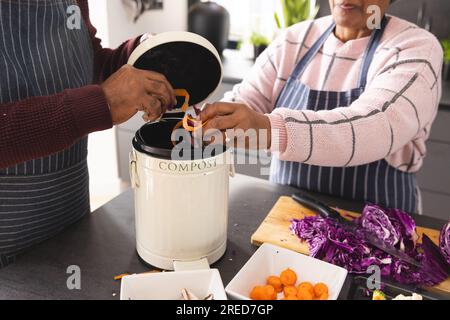 Couple biracial senior portant des tabliers compostant les déchets alimentaires dans la cuisine à la maison Banque D'Images