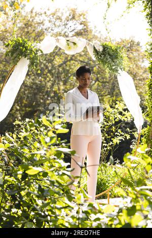 Officiant de mariage féminin biracial focalisé utilisant l'arc de mariage de tablette avec des fleurs dans le jardin ensoleillé Banque D'Images