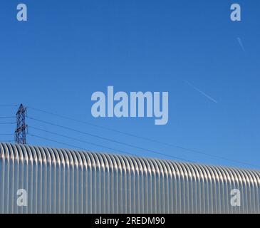 Un pylône électrique sort de derrière un toit en tôle ondulée d'un entrepôt en plein soleil avec un ciel bleu moucheté avec à l'avion tra Banque D'Images