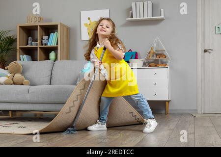 Jolie petite fille balayant le sol sous la moquette avec un balai à la maison Banque D'Images