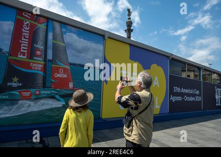 Barcelone, Espagne. 26 juillet 2023. Deux touristes photographient le drapeau du magasin officiel de l'America's Cup Barcelona installé dans le port de Barcelone. Les installations de la compétition de voile American Cup Barcelona commencent à être visibles dans le port de Barcelone. Certaines des bases nautiques des équipes qui participeront au sport de compétition de voile le plus important arborent déjà les drapeaux de leur pays. Certaines équipes s’entraînent même déjà dans les eaux du port de Barcelone. Crédit : SOPA Images Limited/Alamy Live News Banque D'Images