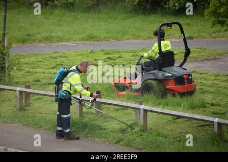 Travailleurs du conseil coupant l'herbe dans l'espace public vu du sentier public, Newbury, Berkshire, Angleterre, Royaume-Uni Banque D'Images