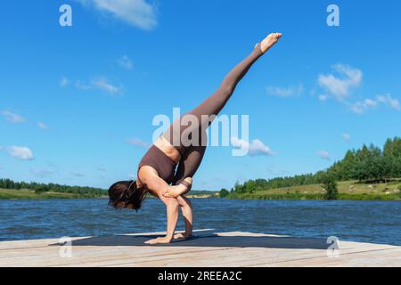 Femme faisant l'exercice Eka Pada Bakasana, pose de grue avec la jambe étendue vers le haut, équilibre asana, support de main pleine longueur, pratique du yoga en vêtements de sport sur le lac Banque D'Images