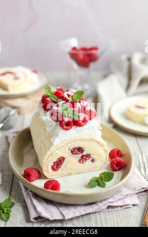 Roulade meringue framboise à la crème vanille, décorée de crème fouettée et de framboises fraîches. Table en bois et fond rose. Banque D'Images