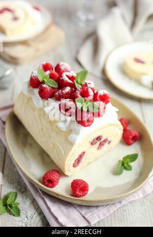 Roulade meringue framboise à la crème vanille, décorée de crème fouettée et de framboises fraîches. Table en bois et fond rose. Banque D'Images