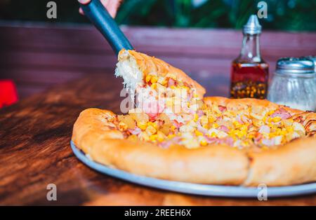 Personne main prenant une tranche de maïs doux et pizza au jambon avec spatule. Délicieuse pizza au jambon avec fromage et maïs sucré Banque D'Images