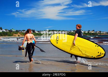 Un jeune homme porte la planche de surf pour sa petite amie alors qu'elle se prépare à attraper une vague sur une planche de paddle à la plage un jour de vacances d'été Banque D'Images