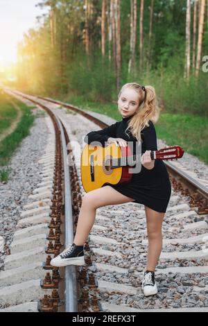 Un adolescent de 13 ans avec une guitare. La fille voyage avec une guitare. La fille est debout sur les rails allant au loin, dans la forêt à Banque D'Images