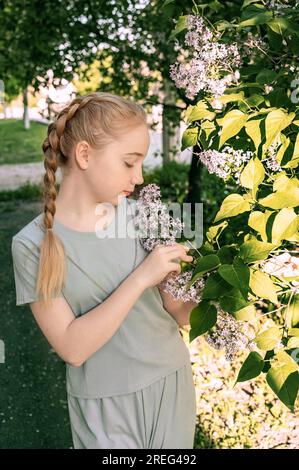 Charmante jeune blonde blonde avec des tresses apprécie l'odeur d'une branche de lilas. Portrait d'une fille de 13-15 ans au printemps Banque D'Images