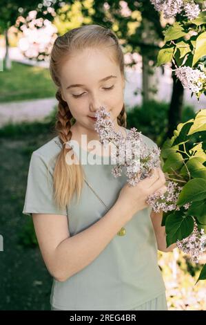 Charmante jeune blonde blonde avec des tresses apprécie l'odeur d'une branche de lilas. Portrait d'une fille de 13-15 ans au printemps Banque D'Images