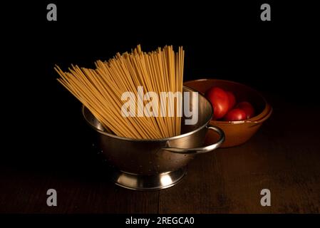 Tomates dans un bol brun et pâtes dans une passoire en métal sur une table en bois sombre avec un éclairage moody. Banque D'Images