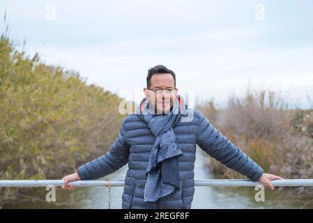 Portrait en longueur de l'homme adulte souriant debout par la balustrade sur le pont tout en regardant la caméra de jour contre la rivière Banque D'Images