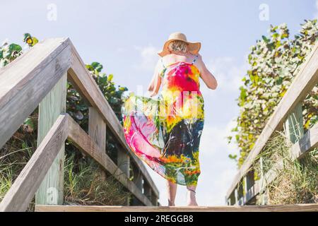 Femme âgée en tie-dye et chapeau de soleil marche vers l'océan sur la promenade Banque D'Images