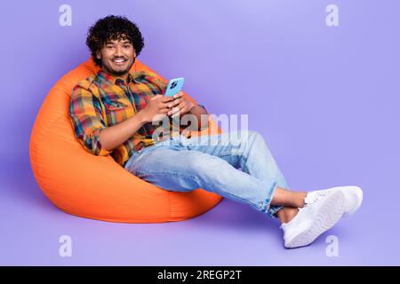 Photo pleine longueur de l'homme optimiste habillé chemise assis dans le pouf orange tenir smartphone lire post isolé sur fond de couleur violet Banque D'Images
