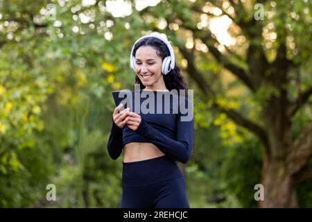 Jeune belle femme latino-américaine marche dans le parc lors d'exercices physiques actifs, femme utilise une application pour écouter des livres audio et de la musique sur le téléphone, souriant sportswoman dans les écouteurs. Banque D'Images