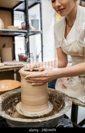 Vue recadrée de potier femelle en tablier créant un vase d'argile sur la roue de poterie dans l'atelier flou Banque D'Images