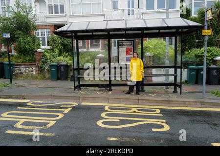 Une femme en manteau mackintosh jaune vif attendant un bus à un arrêt de bus dans le quartier Preston Park de Brighton. 27 juillet 2023 Banque D'Images