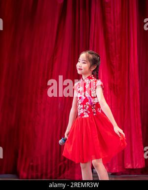 La jeune fille asiatique chante une chanson sur scène à son jour d'activité scolaire, robe de style Qipao, fond rouge rideau Banque D'Images