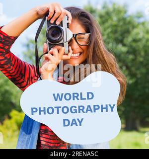 Texte de la journée mondiale de la photographie sur bleu sur femme biracial heureuse utilisant la caméra dans le parc Banque D'Images