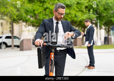 Homme d'affaires attrayant conduisant un scooter de coup de pied au parc de New York. Homme d'affaires dans un costume chevauchant un scooter électrique sur une réunion d'affaires. Âge moyen Banque D'Images