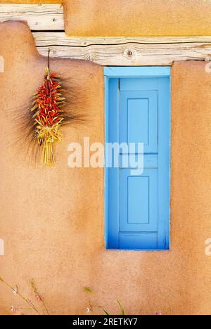 Ranchos de Taos, Nouveau Mexique, États-Unis - 23 juillet 2023 : une istra est accrochée à côté d'un volet en bois peint en bleu sur le mur extérieur d'une maison traditionnelle en adobe. Banque D'Images