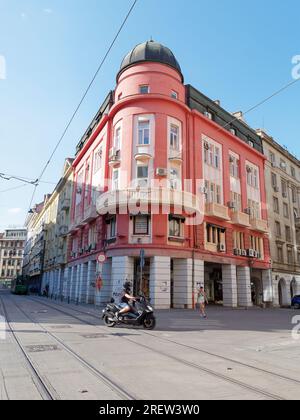 Motocycliste traverse les voies de tramway devant un imposant bâtiment rouge avec dôme au coin de la rue. Sofia, Bulgarie. 29 juillet 2023 Banque D'Images