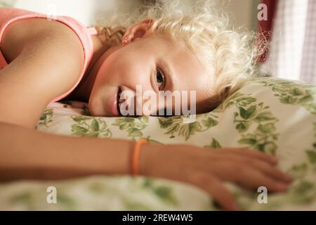 Vue latérale de fille blonde souriante allongée sur le lit et regardant la caméra tout en se relaxant dans la chambre Banque D'Images