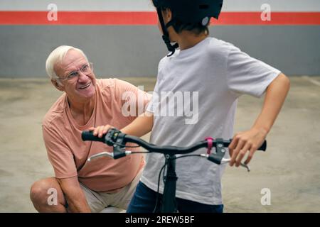 Homme âgé positif en tenue décontractée et lunettes avec garçon anonyme en casque sur vélo passant du temps ensemble sur le parking Banque D'Images