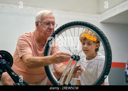 Homme âgé attentif dans les lunettes avec petit-fils mignon réparant roue de vélo moderne tout en passant du temps ensemble sur le parking Banque D'Images