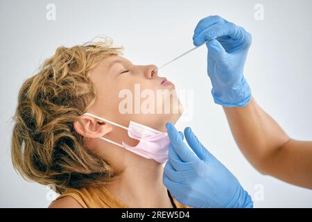 Crop méconnaissable Medic avec écouvillon prenant le test de coronavirus du nez d'un enfant avec les yeux fermés en clinique sur fond clair Banque D'Images