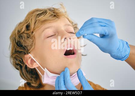 Crop méconnaissable infirmière avec écouvillon prenant le test COVID 19 du nez d'un enfant avec les yeux fermés sur fond clair Banque D'Images