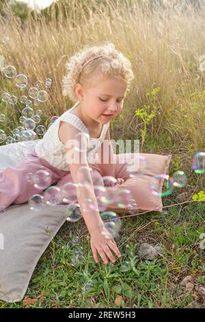 Jolie petite fille en tenue élégante jouant avec des bulles de savon tout en s'asseyant sur des oreillers confortables sur une glade herbacée dans la campagne Banque D'Images