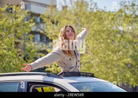 Jeune femme ravie portant un manteau et un rouge à lèvres debout avec les yeux fermés dans le toit ouvrant de la voiture et les bras en élongation dans l'excitation Banque D'Images