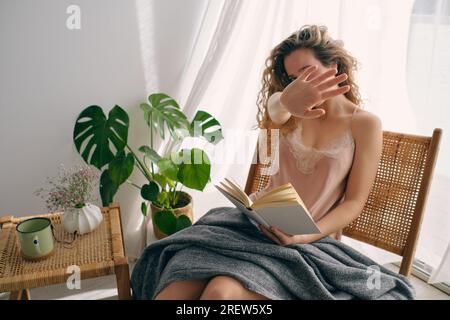 Joyeuse jeune femme en nuisette de soie assise sur une chaise en osier près de la plante monstera et livre de lecture tout en cachant le visage de la caméra dans une chambre confortable avec des lits Banque D'Images