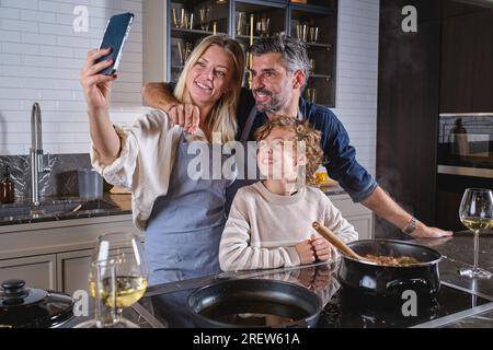 Père hispanique positif non rasé embrassant la femme heureuse et prenant l'autoportrait sur smartphone avec son fils tout en s'amusant ensemble et en cuisinant la nourriture à h. Banque D'Images