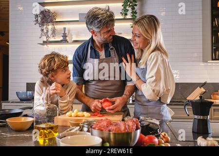 Mari et femme positifs se regardant l'un l'autre tout en épluchant des tomates fraîches près de son fils tout en cuisinant ensemble à table dans la cuisine Banque D'Images