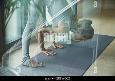 Par verre de femme en forme en vêtements de sport debout sur tapis gris et étirement des jambes dans Prasarita Padottasana Une pose tout en pratiquant le yoga dans la salle de gym Banque D'Images