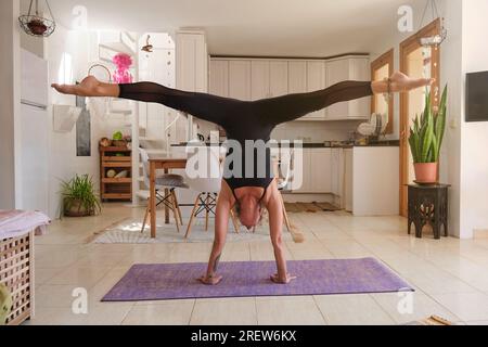 Corps entier de l'athlète féminine en forme en vêtements actifs prenant Adho Mukha Vrikshasana pose et faisant des fentes sur tapis violet tout en pratiquant le yoga dans la cuisine à ho Banque D'Images