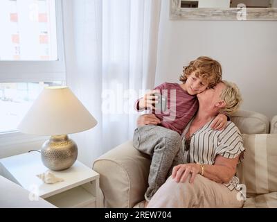 Belle grand-mère embrassant et embrassant petit garçon mignon avec smartphone tout en prenant selfie au salon confortable ensemble Banque D'Images