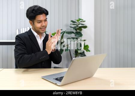 Homme professionnel en tenue d'affaires applaudissant devant l'ordinateur portable tout en vidéoconférence en ligne réunion avec le partenaire d'affaires. Banque D'Images