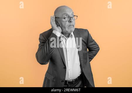 Homme âgé dans la pose d'espionnage sur une conversation. Un homme essayant d'écouter.. La main est mise à l'oreille. Fond orange vif. art Banque D'Images