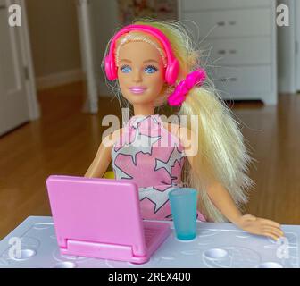 Poupée Barbie en plastique assis à une table avec un ordinateur portable et une tasse écoutant de la musique à travers ses écouteurs. Banque D'Images