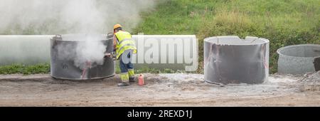 Processus de reconstruction de routes. Un ouvrier installe un collecteur d'eau de pluie. Un homme portant un casque et un respirateur scie un trou dans un tuyau en béton avec une scie Banque D'Images