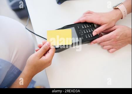 Vue aérienne d'un client consommateur détenant une carte bancaire en plastique doré au-dessus d'un terminal POS ou d'un lecteur de carte de crédit, effectuant des paiements sans espèces sans contact Banque D'Images