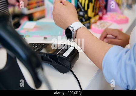 Gros plan d'une cliente utilisant une montre intelligente, effectuant un paiement sans contact à l'aide de la technologie NFC, debout au comptoir dans un magasin avec code à barres sca Banque D'Images