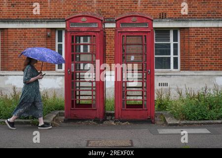 Londres Royaume-Uni. 30 juillet 2023 Un piéton marche avec un parapluie devant des cabines téléphoniques rouges à Wimbledon, au sud-ouest de Londres, pendant des averses de pluie. Crédit amer ghazzal/Alamy Live News Banque D'Images