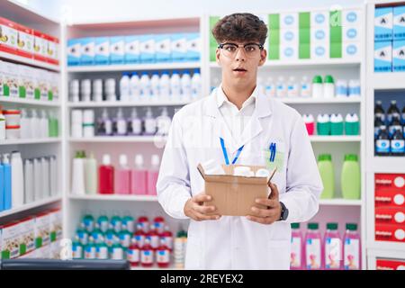 Adolescent hispanique travaillant à la pharmacie pharmacie tenant la boîte avec des pilules effrayé et étonné avec la bouche ouverte pour la surprise, le visage incrédulité Banque D'Images