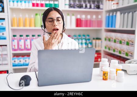 Jeune femme arabe travaillant à la pharmacie pharmacie pharmacie utilisant un ordinateur portable ennuyé bâillant fatigué couvrant la bouche avec la main. agité et somnolence. Banque D'Images