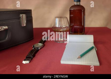 Nature morte rétro - valise, montre-bracelet, whisky et un crayon avec un bloc-notes sur une nappe rouge Banque D'Images