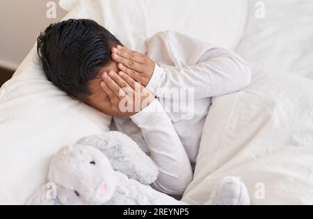 Adorable garçon hispanique couvrant le visage avec les mains couchés sur le lit à la chambre Banque D'Images